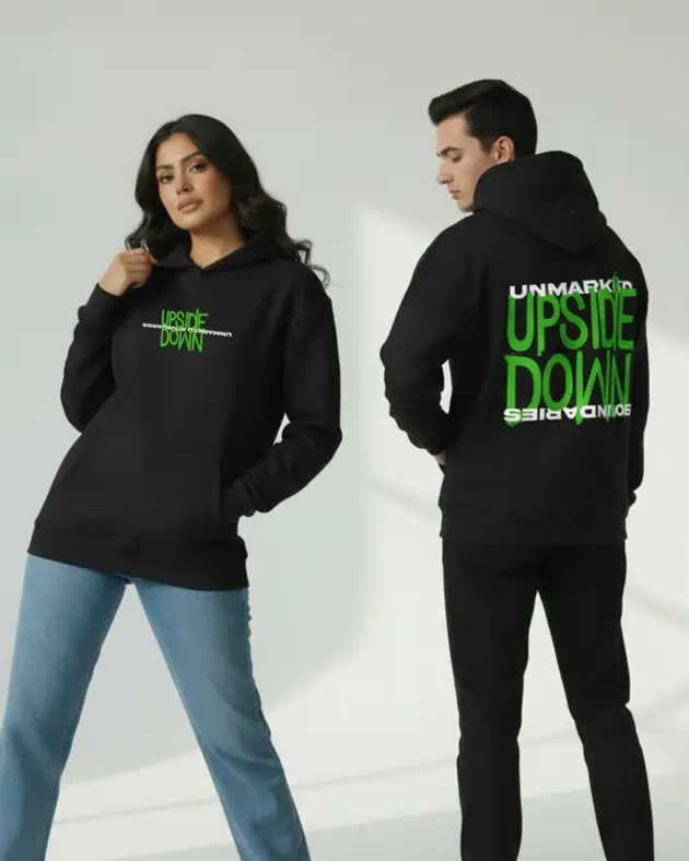 Black oversized unisex hoodie with green typography print worn by male and female models in a minimalist studio setting.
