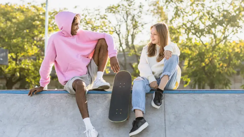 A young man in a pink oversized hoodie and a young woman in a cream hoodie sitting on a concrete skate ramp, smiling at each other with a skateboard between them.