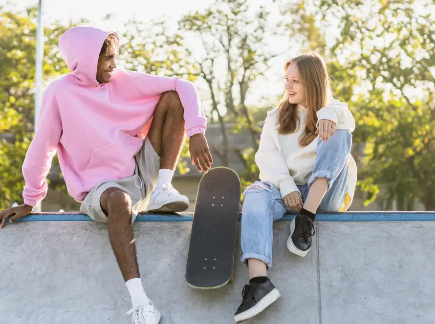 A young man in a pink oversized hoodie and a young woman in a cream hoodie sitting on a concrete skate ramp, smiling at each other with a skateboard between them.