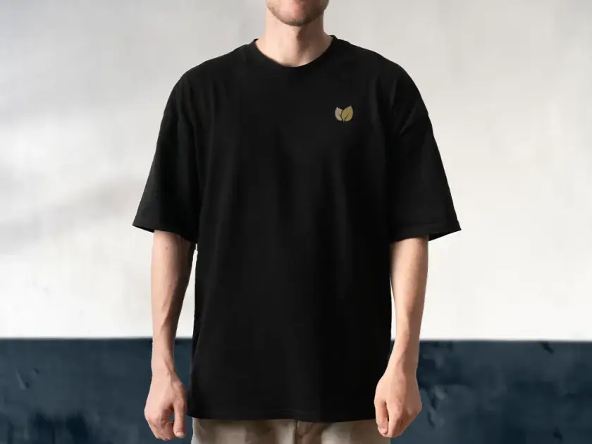 A man wearing a black oversized t-shirt with a small gold leaf logo on the left chest, standing against a neutral textured wall.