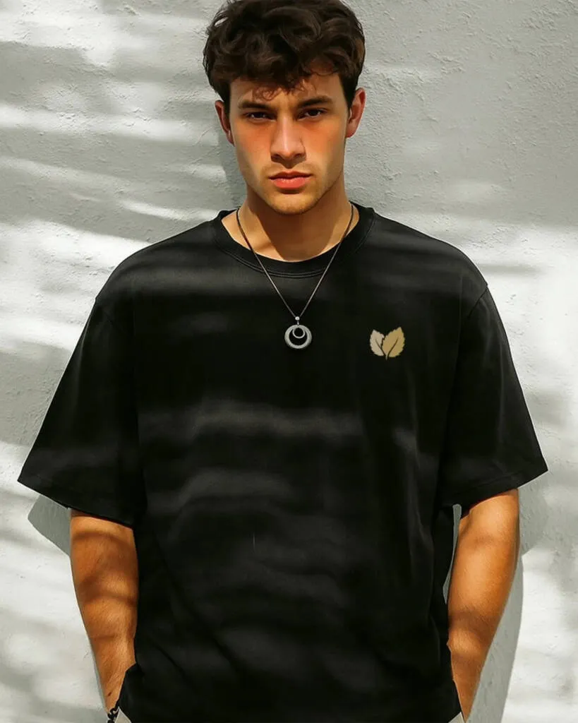 A man wearing a black oversized t-shirt with a small gold leaf logo and a circular pendant necklace, standing against a sunlit white wall with tree shadows.
