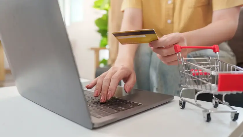 Close-up of a person holding a credit card while typing on a laptop next to a miniature red shopping cart on a white desk.