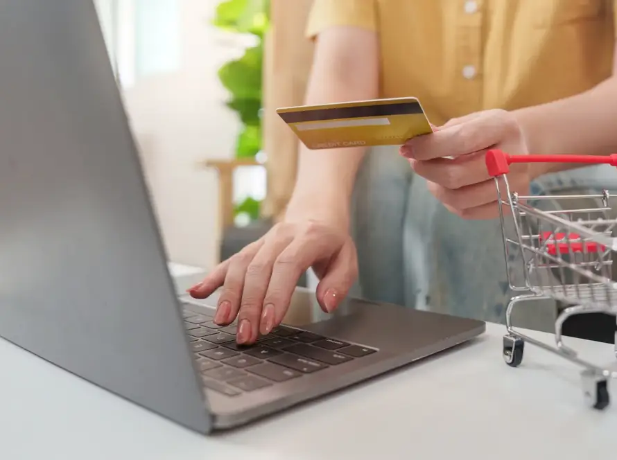 Close-up of a person holding a credit card while typing on a laptop next to a miniature red shopping cart on a white desk.