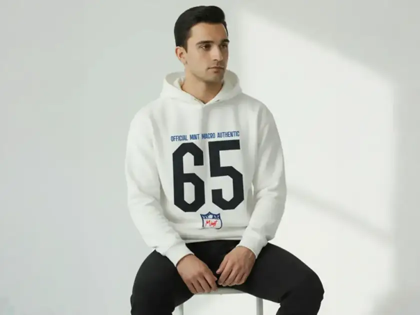 A man sitting on a stool wearing a white Mint Macro hoodie with a large black "65" jersey-style print and navy blue branding.