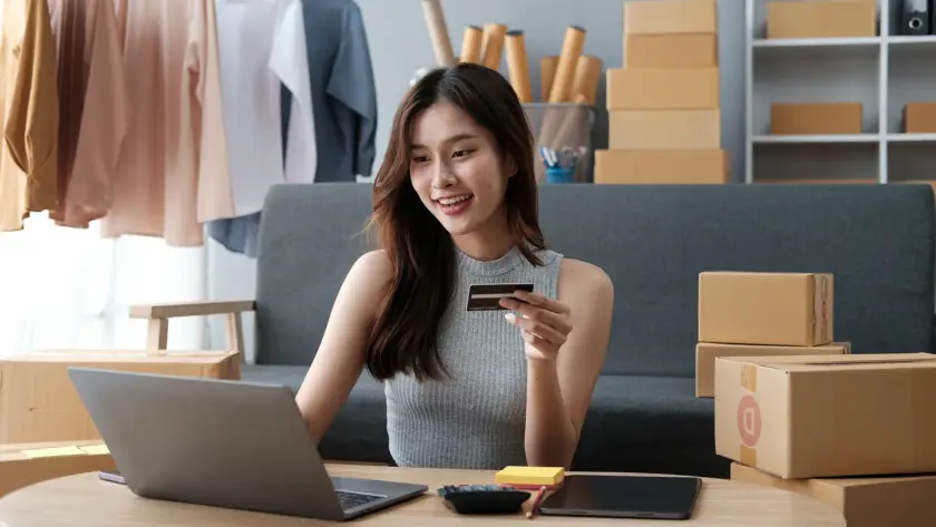 A smiling woman sitting at a desk with a laptop and credit card, surrounded by shipping boxes and a clothing rack in a bright home office.