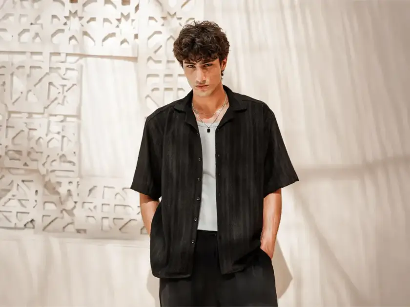 A man wearing an open black textured short-sleeve shirt over a white undershirt and silver necklaces against a patterned white wall.