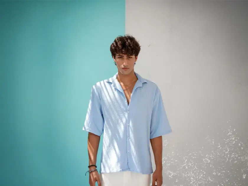 A man wearing a light blue textured button-down shirt and white trousers standing against a split teal and white wall.