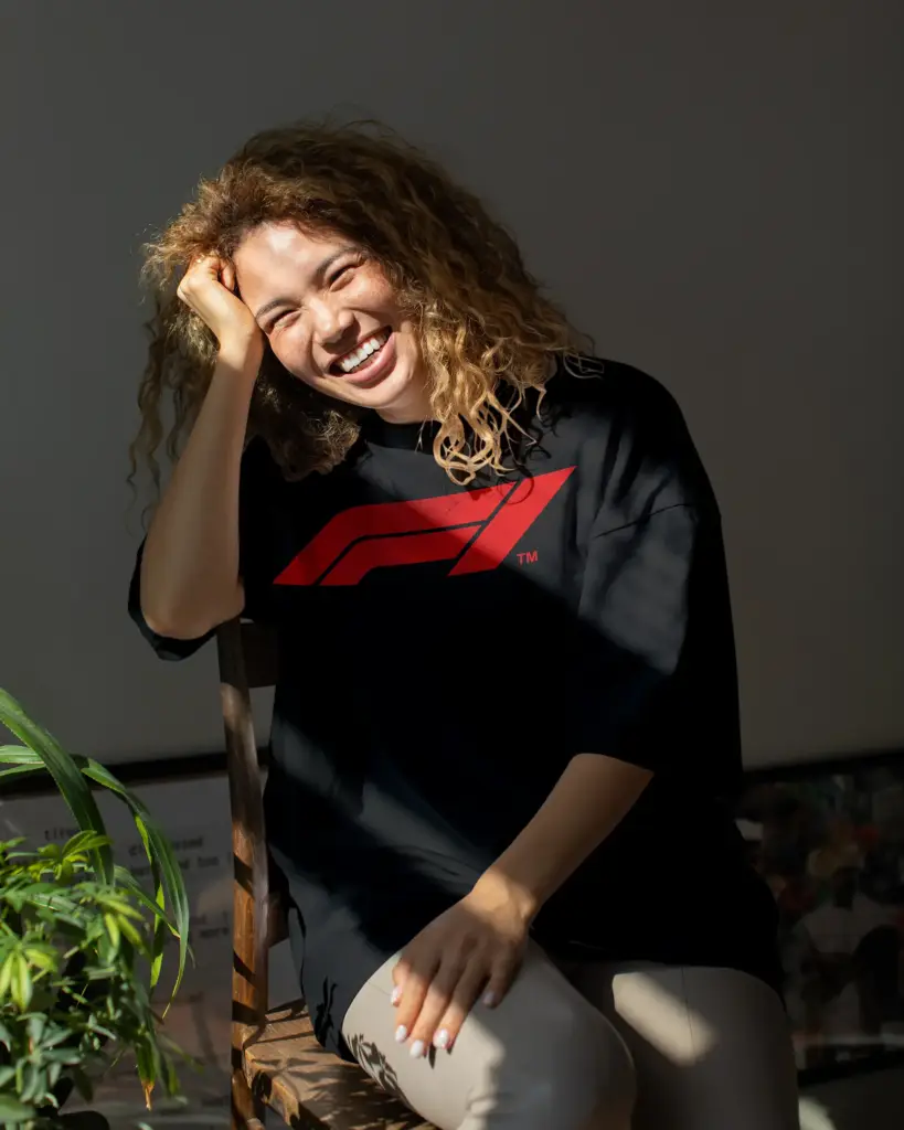 A smiling woman with curly hair wearing a Mint Macro black oversized t-shirt with a red F1 logo, sitting in natural sunlight.