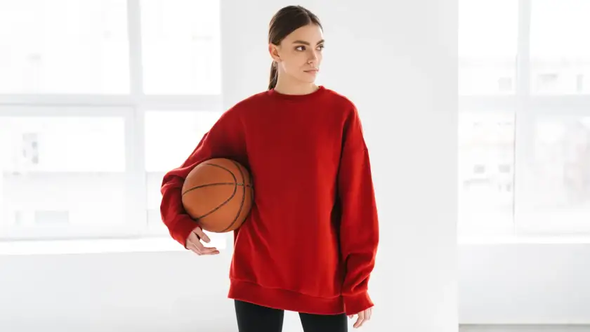 A woman in a red oversized sweatshirt holding a basketball under her arm, standing in a bright, white studio.