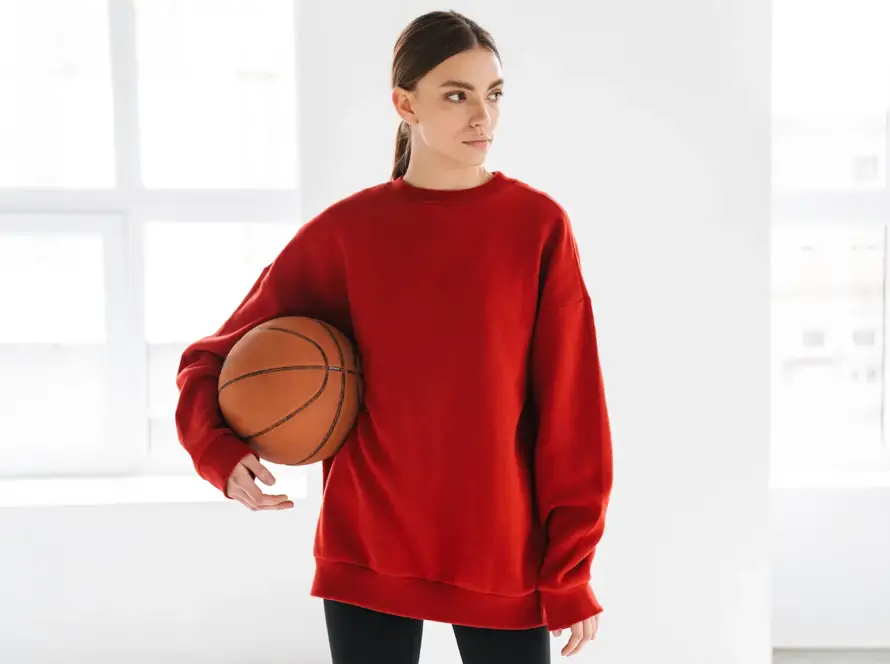 A woman in a red oversized sweatshirt holding a basketball under her arm, standing in a bright, white studio.