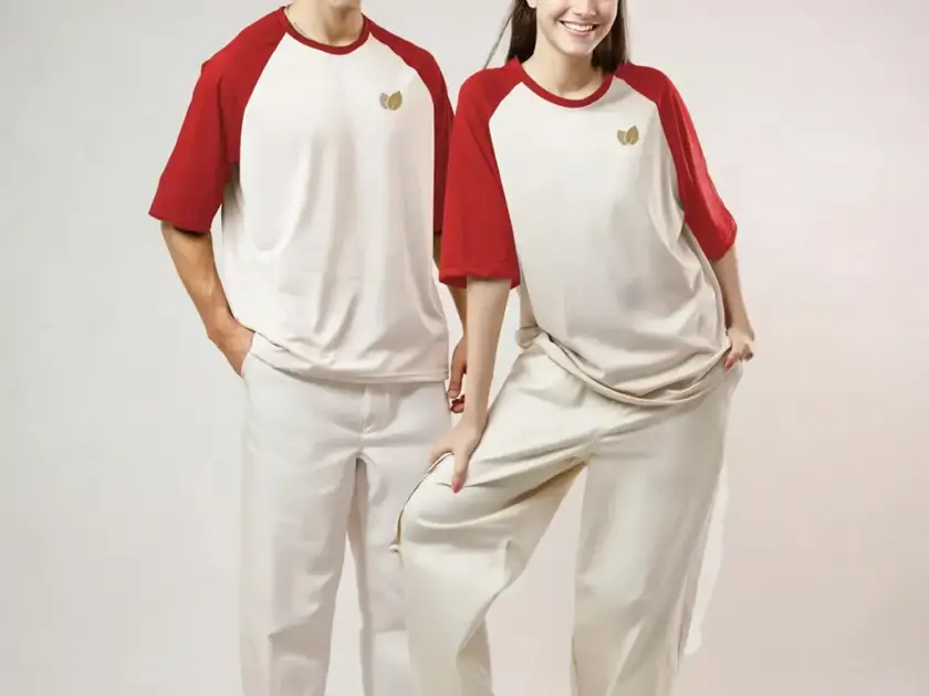 A man and a woman modeling matching cream-colored raglan t-shirts with red sleeves and small gold leaf logos, paired with light-colored trousers.