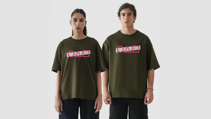 A man and a woman modeling olive green oversized t-shirts with a pink circle, triangle, and square graphic on a white bar, paired with black pants.