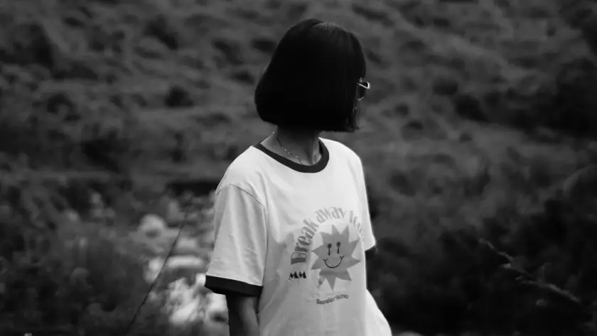A woman with a short bob haircut wearing a white Mint Macro ringer tee with "Break Away" and a sun graphic, standing in a blurred outdoor landscape.