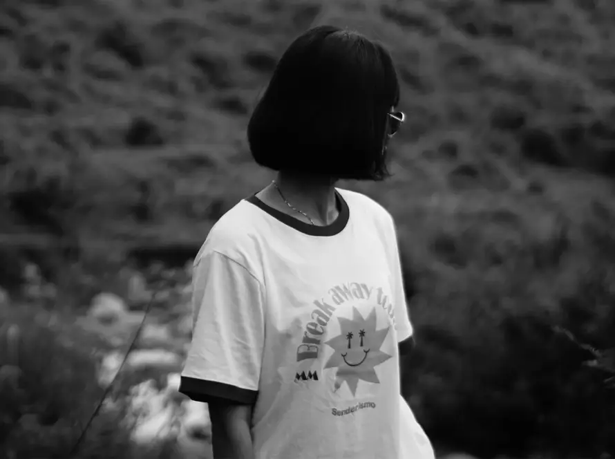 A woman with a short bob haircut wearing a white Mint Macro ringer tee with "Break Away" and a sun graphic, standing in a blurred outdoor landscape.