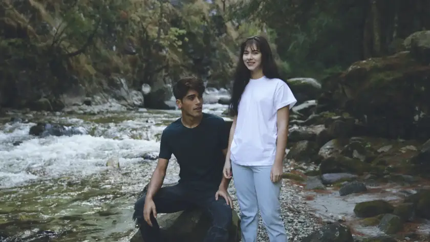 A man in a black t-shirt and a woman in a white t-shirt posing by a rocky river in a lush forest setting.