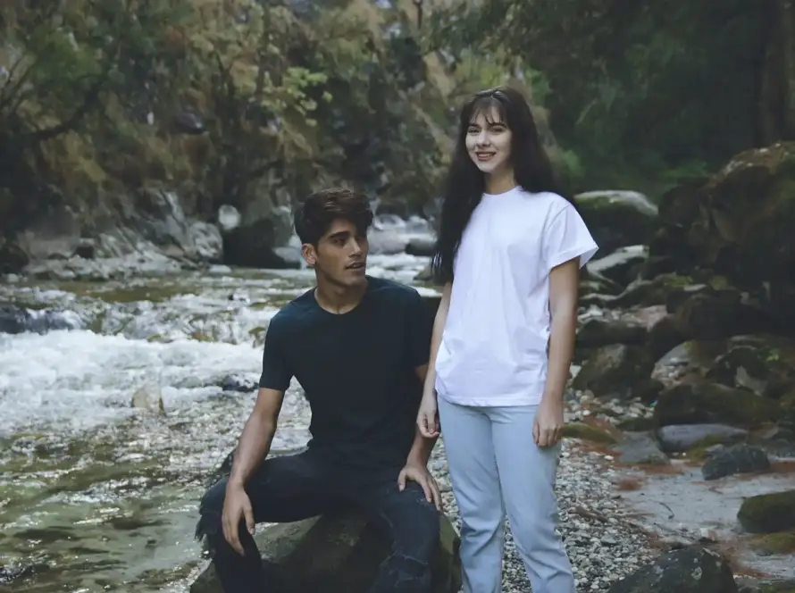 A man in a black t-shirt and a woman in a white t-shirt posing by a rocky river in a lush forest setting.