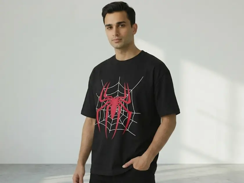 A male model in a minimalist studio setting wearing a black oversized t-shirt featuring a large red spider and white web graphic.