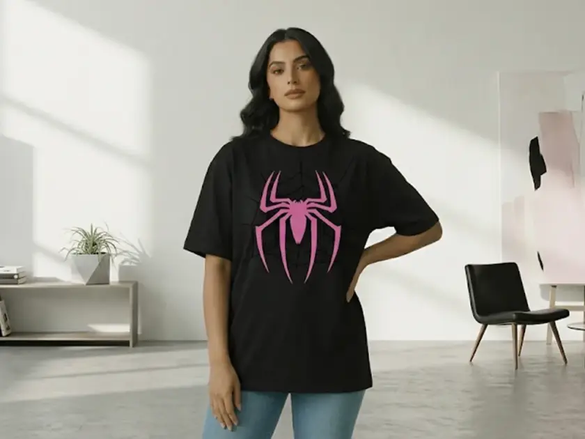 A woman posing in a black oversized t-shirt featuring a large pink spider and web graphic on the front, set in a bright, modern interior.