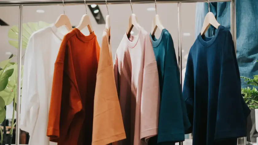 row of high-quality oversized t-shirts in earthy tones—white, burnt orange, tan, dusty pink, teal, and navy blue—hanging on wooden hangers on a sleek metal clothing rack in a modern retail store.