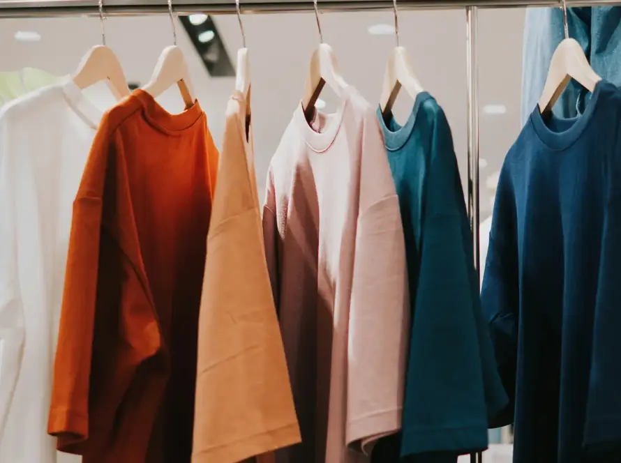 row of high-quality oversized t-shirts in earthy tones—white, burnt orange, tan, dusty pink, teal, and navy blue—hanging on wooden hangers on a sleek metal clothing rack in a modern retail store.