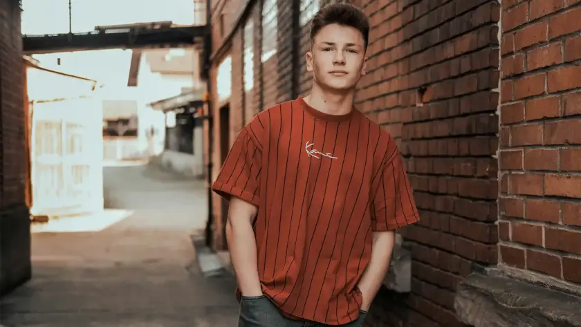 A young man wearing an oversized burnt orange pinstripe t-shirt with script embroidery, standing in an urban alleyway in Pakistan.