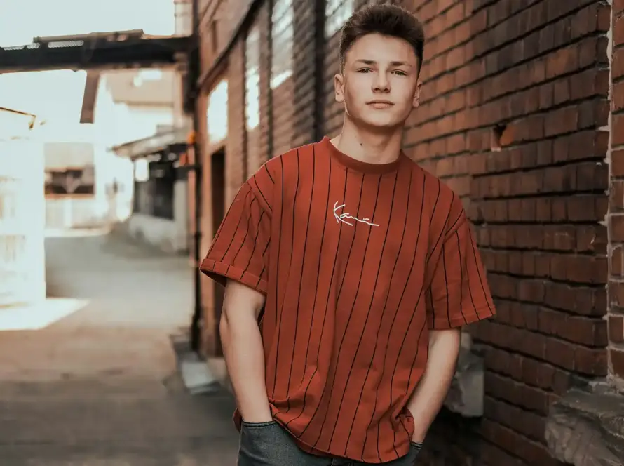 A young man wearing an oversized burnt orange pinstripe t-shirt with script embroidery, standing in an urban alleyway in Pakistan.