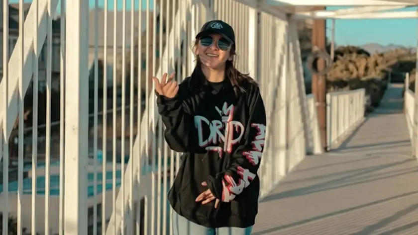 A person wearing a black oversized "DRIP" graphic hoodie, sunglasses, and a baseball cap posing on a white bridge during sunset.