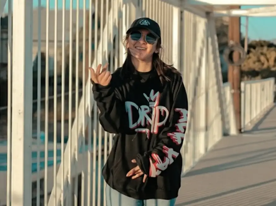 A person wearing a black oversized "DRIP" graphic hoodie, sunglasses, and a baseball cap posing on a white bridge during sunset.