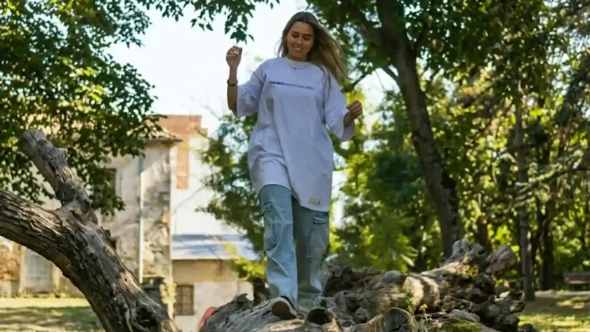 A young woman smiling and walking across a fallen tree trunk in a park, wearing an oversized white graphic t-shirt and light-wash cargo jeans in a landscape orientation.