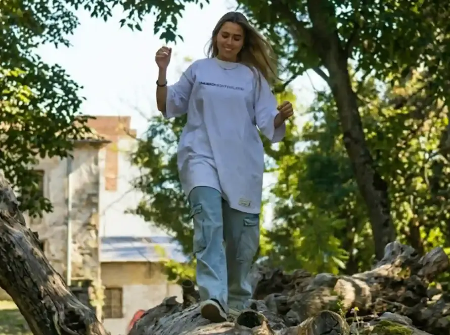 A young woman smiling and walking across a fallen tree trunk in a park, wearing an oversized white graphic t-shirt and light-wash cargo jeans in a landscape orientation.