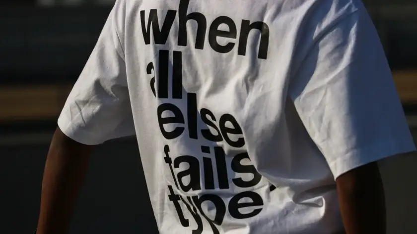 A close-up shot of the back of a person wearing a white oversized streetwear t-shirt featuring bold, black sans-serif typography that reads "when all else fails type." The shirt has a heavy cotton texture and a relaxed drop-shoulder fit.