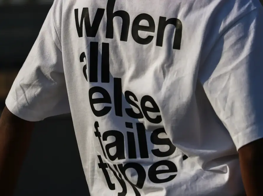A close-up shot of the back of a person wearing a white oversized streetwear t-shirt featuring bold, black sans-serif typography that reads "when all else fails type." The shirt has a heavy cotton texture and a relaxed drop-shoulder fit.