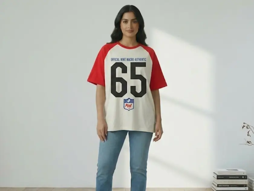 A woman wearing an oversized white and red raglan t-shirt featuring the "Official Mint Macro Authentic 65" graphic, paired with blue jeans in a bright, minimalist studio