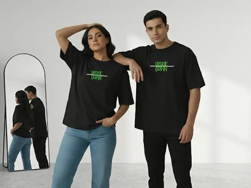 A medium shot of a teenage male model with dark hair, looking directly at the camera, leaning against a weathered red brick wall in an industrial alleyway. He is wearing a dark orange, vertically pinstriped short-sleeve t-shirt with a white script logo that reads 'Kana' on the left chest. His hands are in the front pockets of dark grey pants. The alley behind him is slightly out of focus, showing industrial buildings and a bright end-of-alley opening under a clear sky.