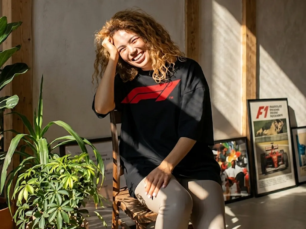 A woman with curly hair laughing while wearing a black oversized t-shirt with a red F1 logo, sitting on a wooden chair in a sunlit room with plants.