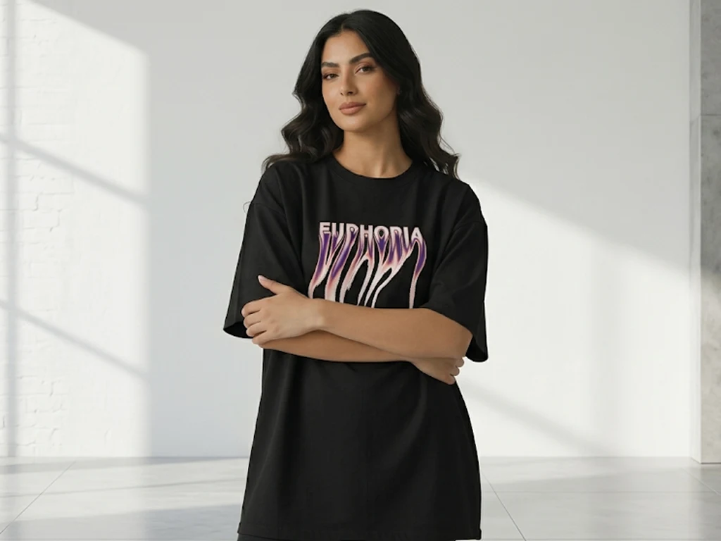 A woman standing in a bright, minimalist studio wearing a black oversized t-shirt. The shirt features a central graphic with the word "EUPHORIA" in a distorted, melting typography style with purple and pink gradients.