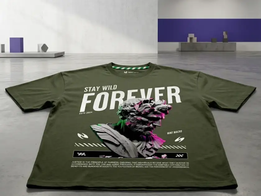 An olive green oversized t-shirt laid flat on a polished concrete floor in a minimalist studio setting. The shirt features a large graphic of a classical bust with neon pink and green accents, the text "STAY WILD FOREVER," and the brand name "MINT MACRO."
