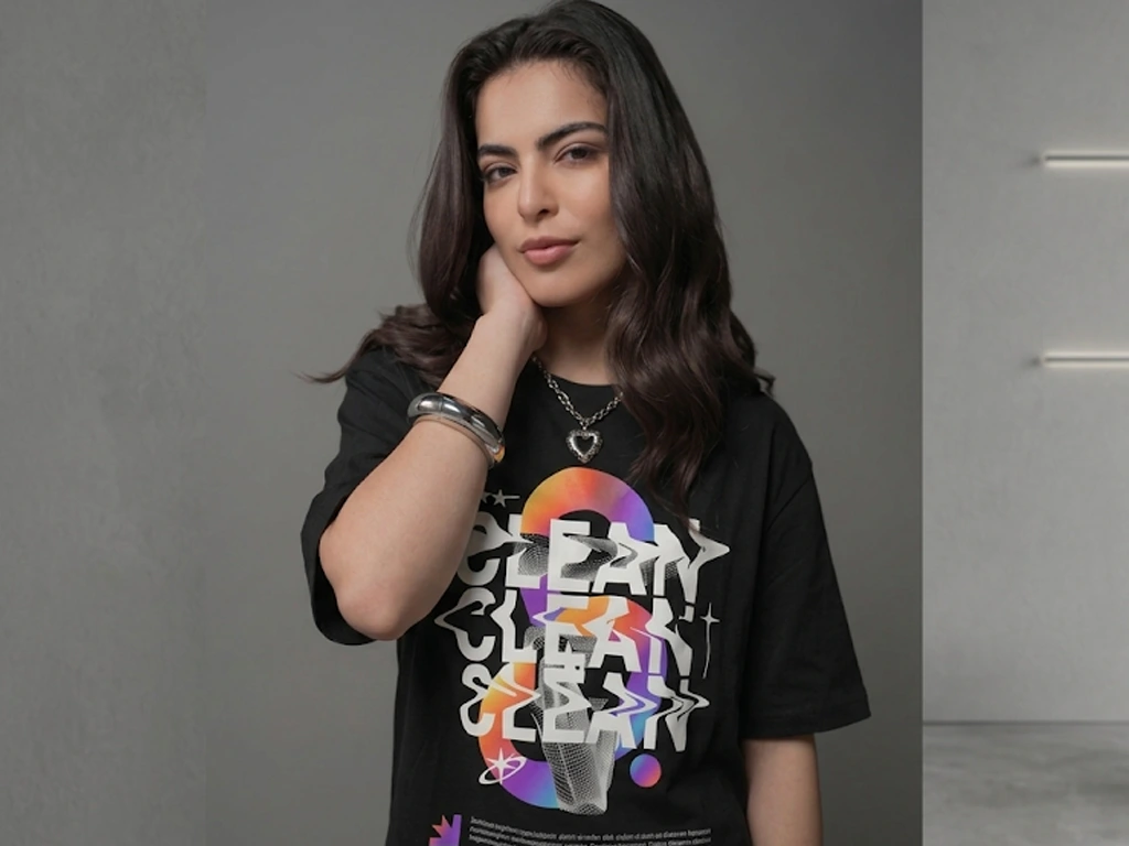A woman with long wavy hair poses in a landscape-oriented shot, wearing a black t-shirt with large, colorful "CLEAN" graphic text and a silver heart pendant necklace.