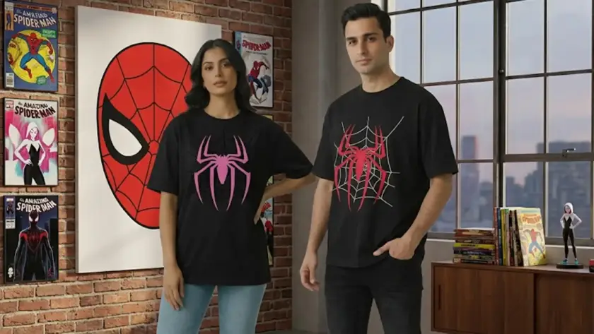 A man and woman standing in a loft-style room with brick walls, wearing black oversized t-shirts featuring pink and red spider web graphics, surrounded by Spider-Man comic posters and collectibles