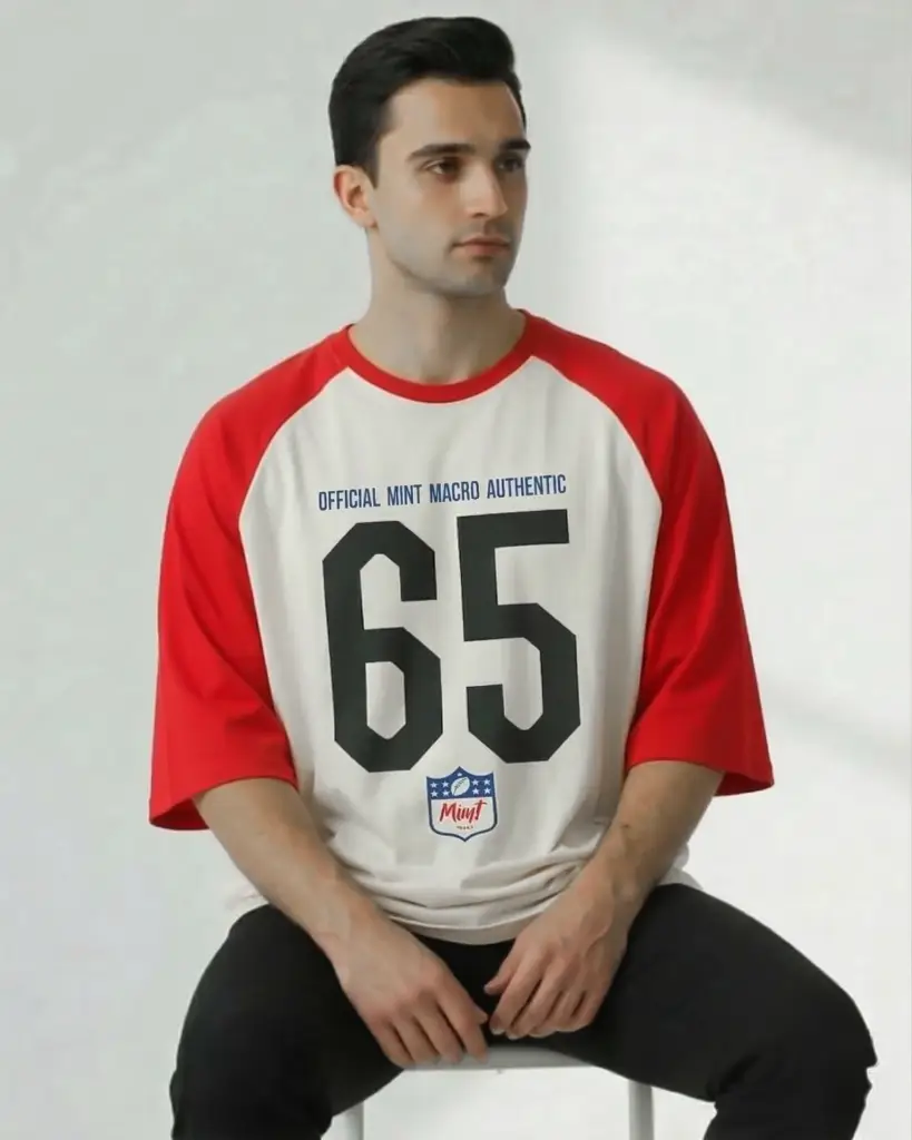 A man sitting against a neutral background wearing an oversized raglan-style t-shirt. The shirt features cream-colored body fabric with contrasting bright red sleeves. The front displays a large black "65" graphic, the text "OFFICIAL MINT MACRO AUTHENTIC" in blue, and a small sports-style shield logo at the bottom.