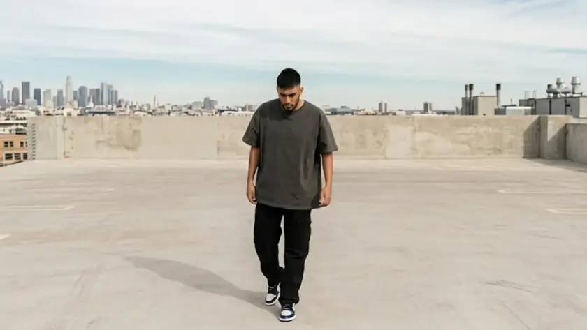 A man in a loose-fitting dark grey t-shirt and black cargo pants walks across an open concrete rooftop parking lot with a city skyline visible in the background.