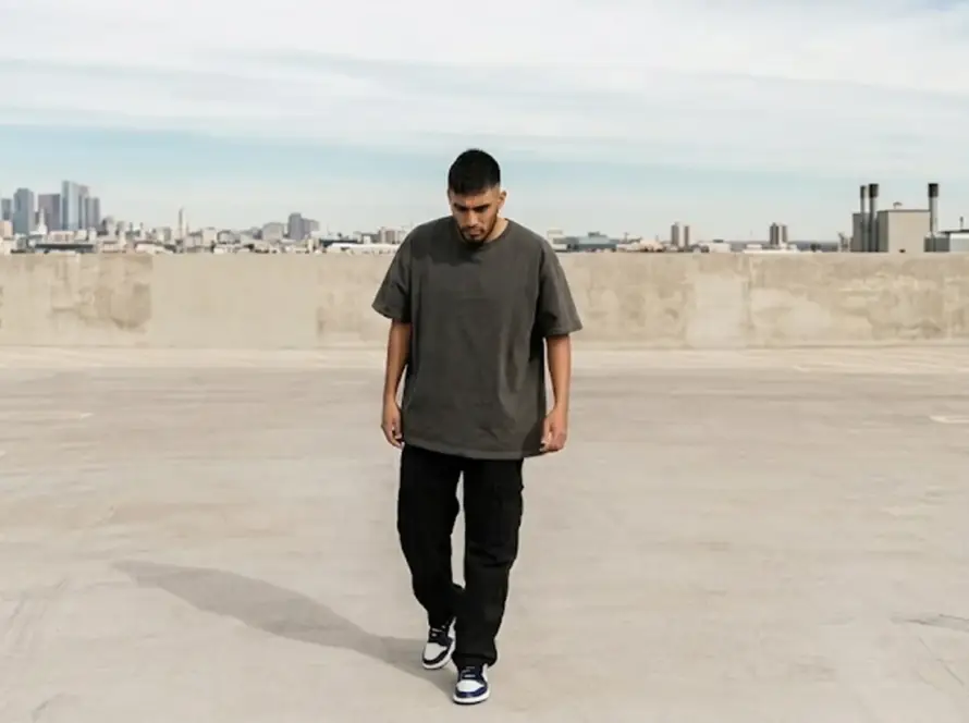 A man in a loose-fitting dark grey t-shirt and black cargo pants walks across an open concrete rooftop parking lot with a city skyline visible in the background.
