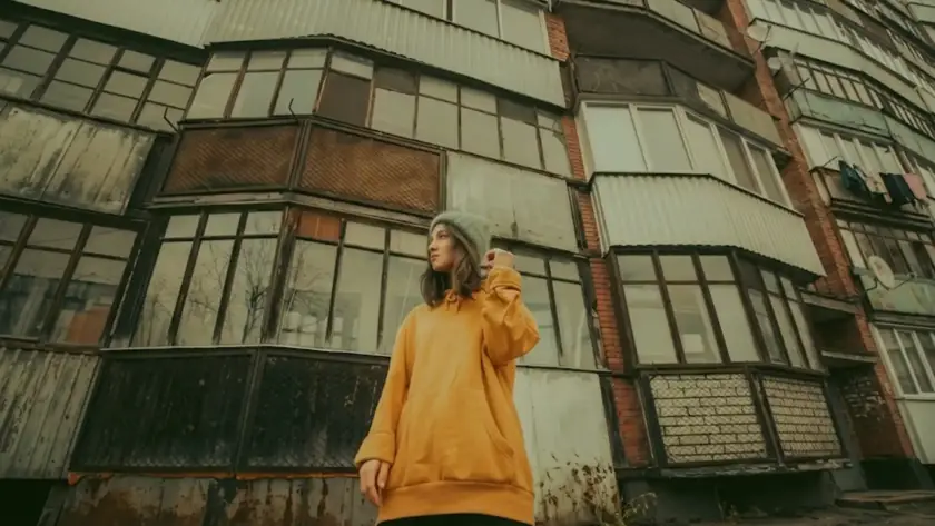 A young person in an orange hoodie and light green beanie stands in front of a multi-story, weathered residential building with many balconies, taken from a low angle.