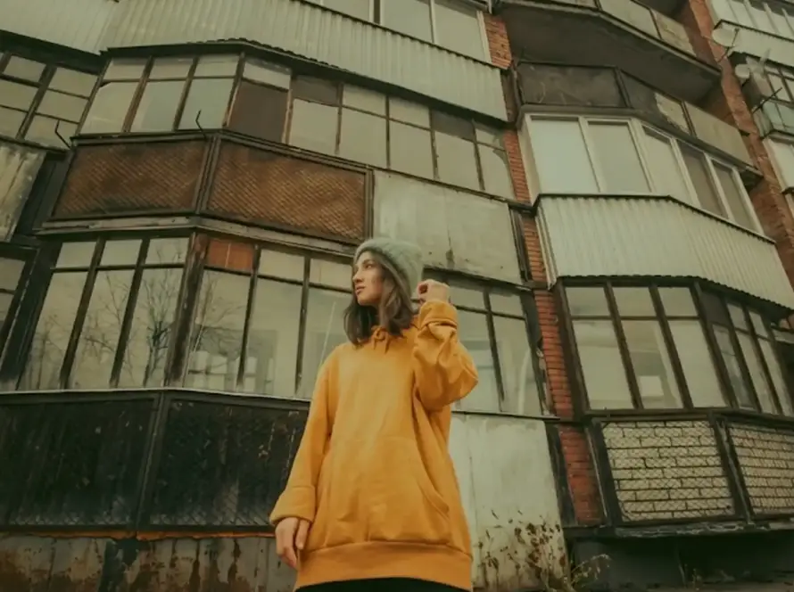 A young person in an orange hoodie and light green beanie stands in front of a multi-story, weathered residential building with many balconies, taken from a low angle.