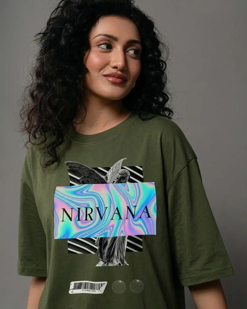 Close-up of a woman with curly hair modeling Mint Macro's olive green oversized t-shirt, featuring a front graphic of a classical angel statue overlaid with a wavy iridescent holographic "NIRVANA" banner against a grey studio background.