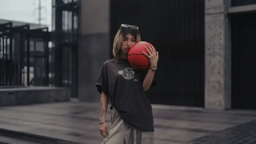 A young woman with blonde-highlighted hair wearing a loose-fitting dark graphic t-shirt and light cargo pants, holding a red basketball near an urban building structure.
