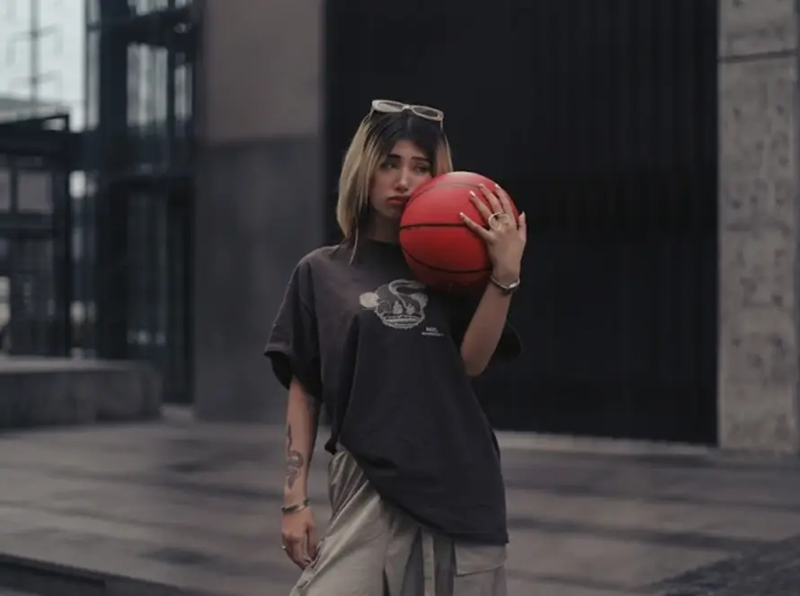 A young woman with blonde-highlighted hair wearing a loose-fitting dark graphic t-shirt and light cargo pants, holding a red basketball near an urban building structure.