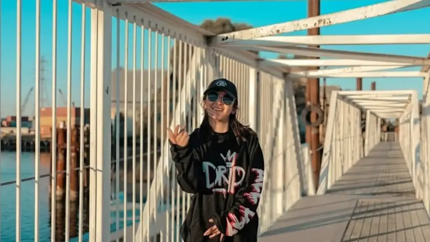 A wide landscape photograph of a person smiling while posing on a white industrial metal bridge walkway. The individual is wearing an oversized black hoodie with bold graphic text, light blue jeans, a black cap, and round sunglasses. The background features a calm blue waterway and an industrial harbor with shipping containers and cranes under a clear sky.