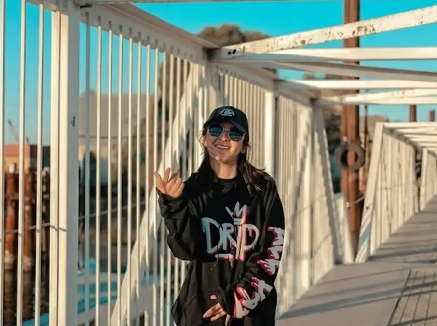 A wide landscape photograph of a person smiling while posing on a white industrial metal bridge walkway. The individual is wearing an oversized black hoodie with bold graphic text, light blue jeans, a black cap, and round sunglasses. The background features a calm blue waterway and an industrial harbor with shipping containers and cranes under a clear sky.