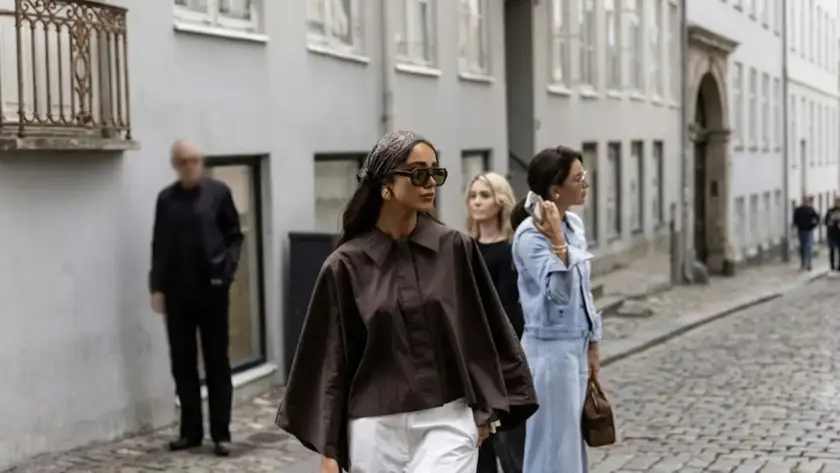 A woman in a brown wide-sleeve shirt, white trousers, and a patterned headscarf walking on a cobblestone European street during a fashion event.